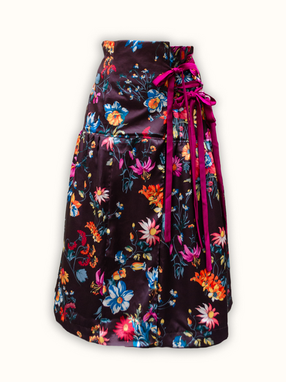 A high-waisted floral skirt made from deadstock fabric, featuring vibrant multicolored flowers like pink, blue, yellow, and orange blooms. The satin-like, slow fashion design includes a tiered structure and ribbon laces tied on the side, reflecting a colorful, made-to-order aesthetic